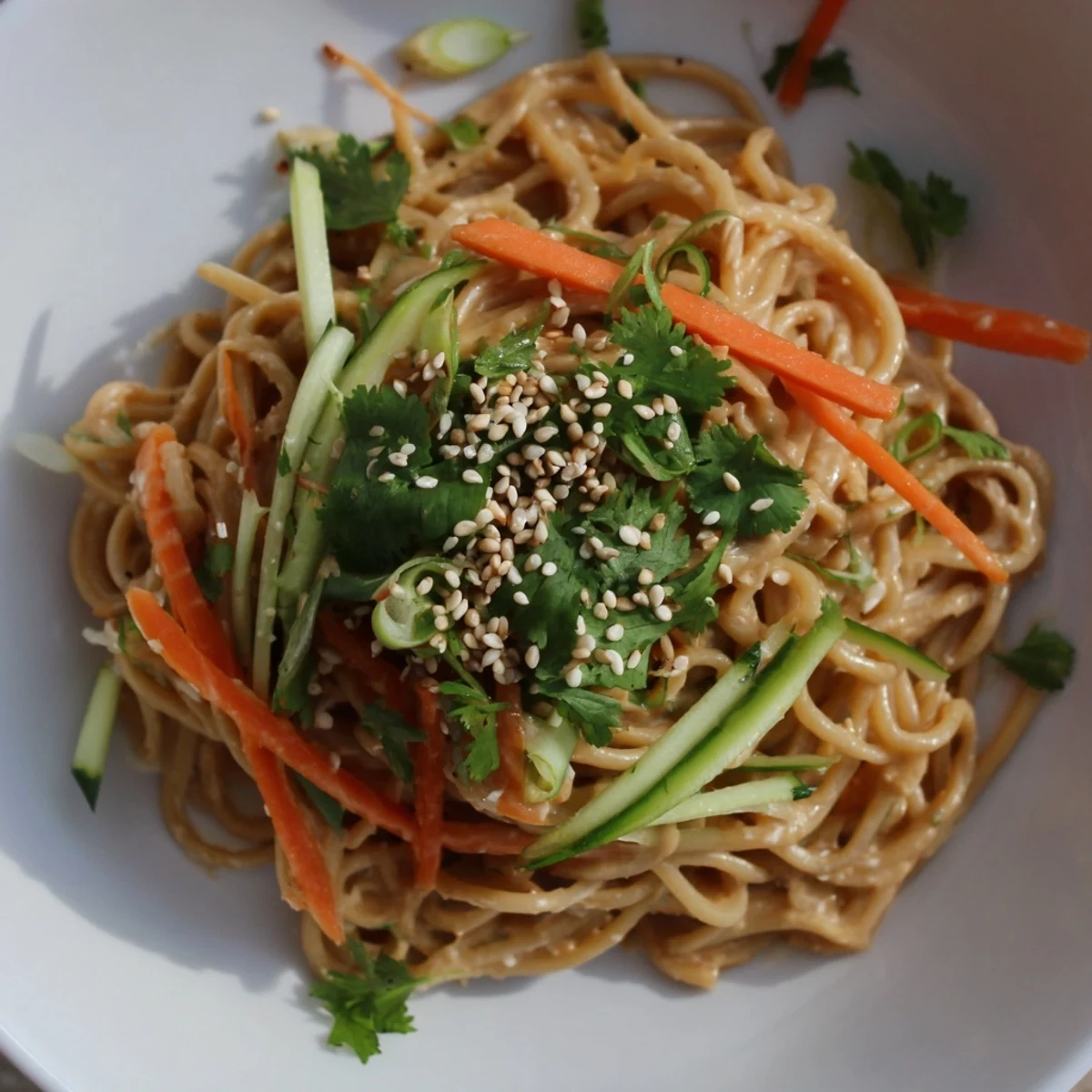 A vibrant bowl of cold sesame noodles, topped with colorful carrots, cucumbers, and fresh cilantro.