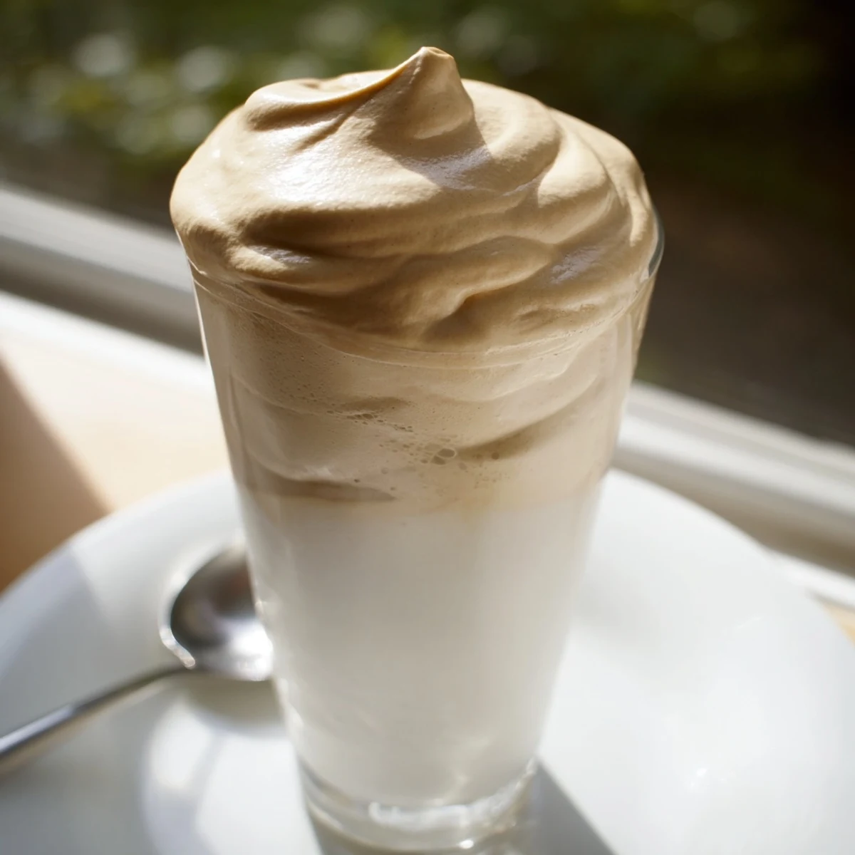 Fluffy, light-brown whipped Dalgona coffee is served in a tall glass filled with milk and ice.