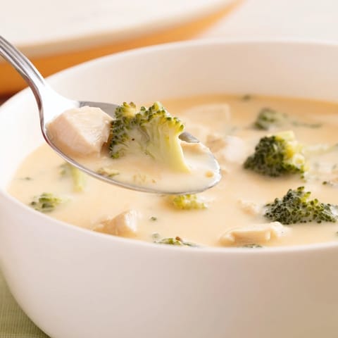 A spoon dips into rich, velvety Chicken Broccoli Cheddar Soup garnished with extra cheese, ready to serve with crusty bread.